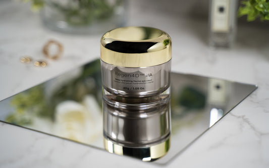 Regen4D® eye cream placed on a reflective metal surface, with elegant jewelry and a lush green shrub in the background. The white flowers are reflected in the metal surface, adding a touch of beauty to the image.